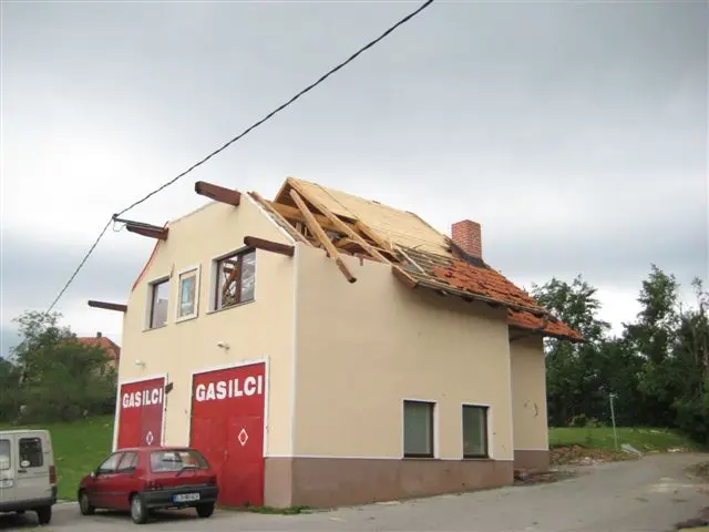 Manjša gasilska stavba s poškodovano streho in dvema avtomobiloma pred objektom.